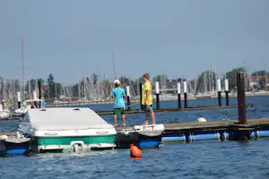 Bootsliegeplatz am Hafen: zwei Personen laufen am Steg entlang, neben einem kleinen Boot und einem orangefarbenen Schwimmkörper.