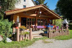 Holzhütte mit überdachter Terrasse, Sitzgruppe und Tisch, Blumenbeete im Garten sowie Grill am Rand.