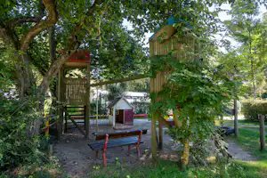 Spielplatz mit Baumhaus und Schaukeln, umgeben von Bäumen und Grünpflanzen, im Hintergrund weitere kleine Gebäude.