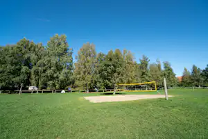 Volleyballfeld auf grünem Rasen mit gelbem Netz, Sandfläche und hohen Bäumen am Campingplatz.