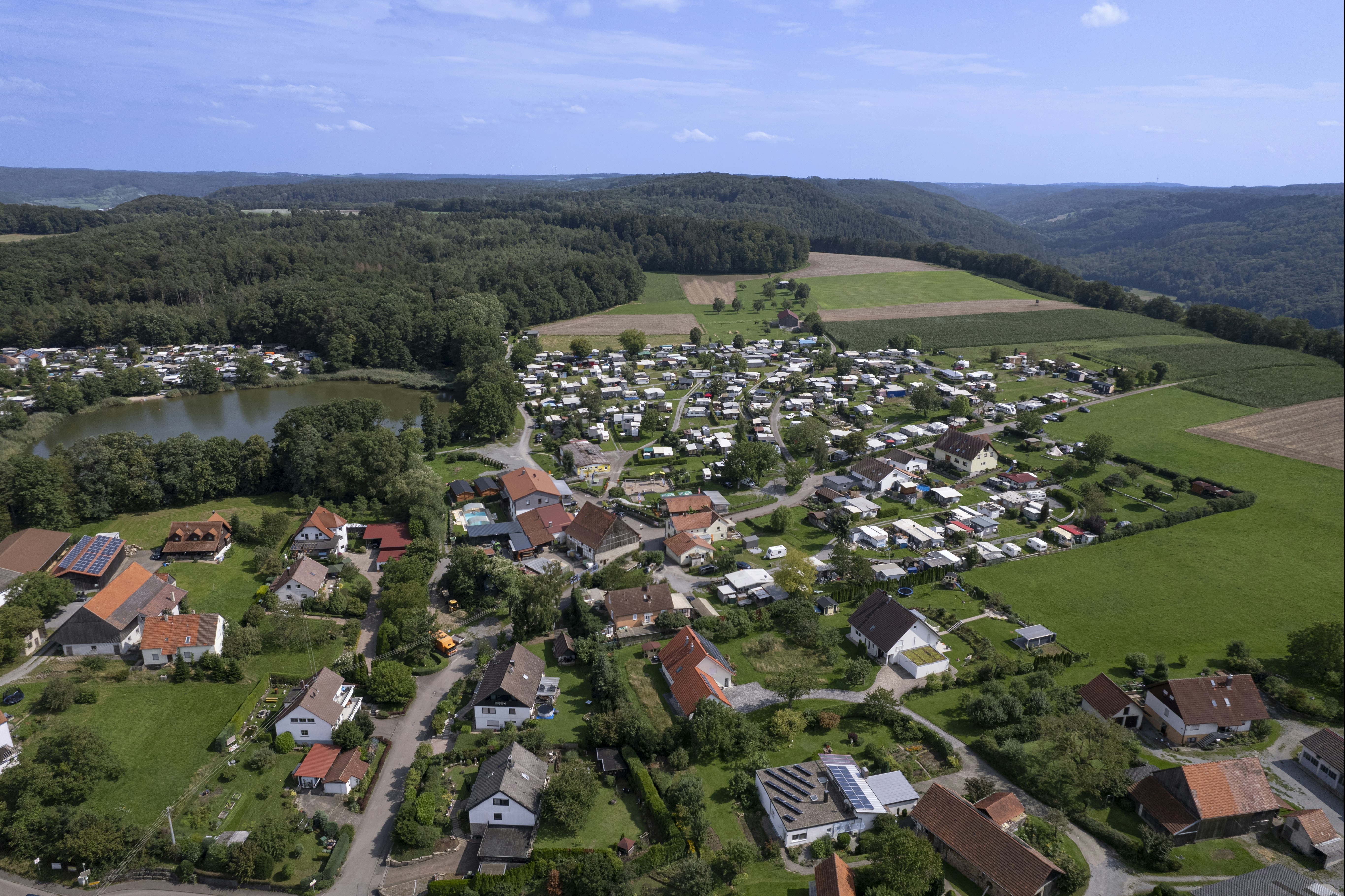 Luftaufnahme eines Campingplatzes mit vielen Wohnwagen und Zelten, umgeben von Wiesen, Feldern und Waldhängen.