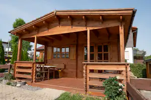 Holzveranda mit überdachter Terrasse, Holzhütte und Sitzbank, davor gepflasterter Weg und Hecken im Garten.