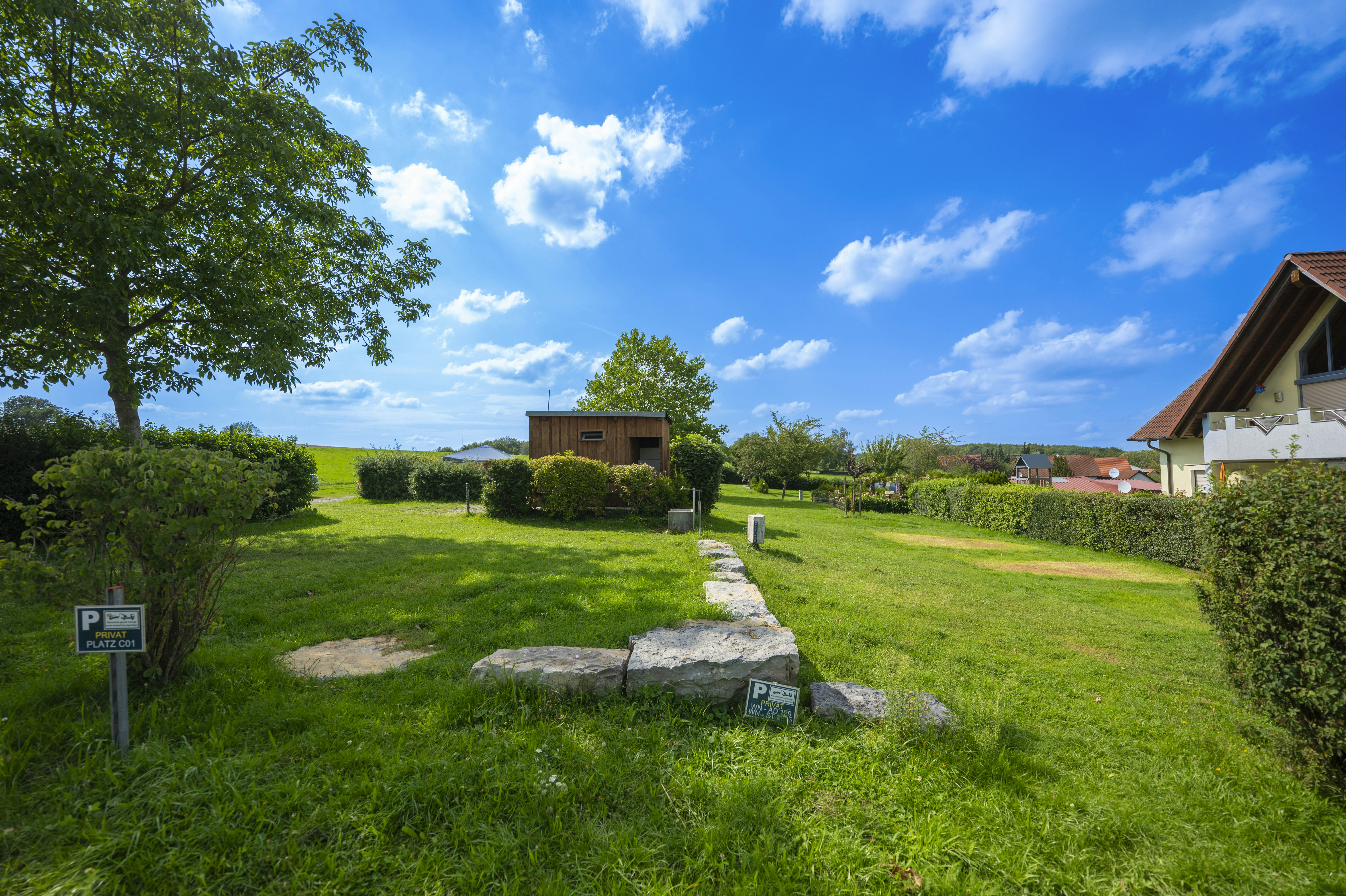 Campingwiese mit gepflastertem Weg, kleinen Gebäuden und Bäumen, im Hintergrund ein Haus und grünes Gelände.
