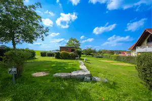 Campingwiese mit gepflastertem Weg, kleinen Gebäuden und Bäumen, im Hintergrund ein Haus und grünes Gelände.
