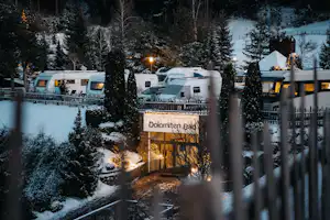 Campingplatz im Winter mit verschneiten Wohnwagen, beleuchtetem Eingang und Blick auf Bäume und Gebäude im Hintergrund.