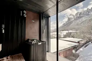 Balkon an einem Holzhaus mit Geländer, Blick ins Tal und auf verschneite Berge, daneben ein Grill und Sitzbank.