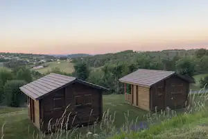 Zwei Holz-Hütten auf einem Campingplatz am Hang, mit Gras, Bäumen und Blick ins Tal bei Dämmerung.