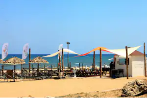 Strand-Camping mit Holzterrasse, Sonnenschirmen und bunten Segeltüchern am Meer, Liegestühle und Tische im Vordergrund.