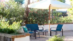 Sitzgruppe mit zwei Campingstühlen unter einem Sonnenschirm auf einer Terrasse, umgeben von Blumenbeeten und Bäumen.