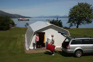 Campingzelt auf einer Wiese am See, daneben ein Auto und zwei Personen vor dem Eingang, im Hintergrund Berge und Wasser.
