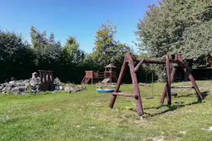 Spielplatz im Campinggarten: Holzschaukel, Klettergerüst und Sandkasten auf Rasen, mit Bäumen und Steinmauer im Hintergrund.
