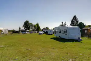 Wohnmobile auf grünem Campingplatz, daneben Autos und Bäume, im Hintergrund weitere Fahrzeuge und ein Gebäude.