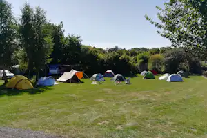 Zeltwiese mit mehreren Campingzelten, Campingstühlen und einem Wohnwagen im Hintergrund, umgeben von Bäumen.