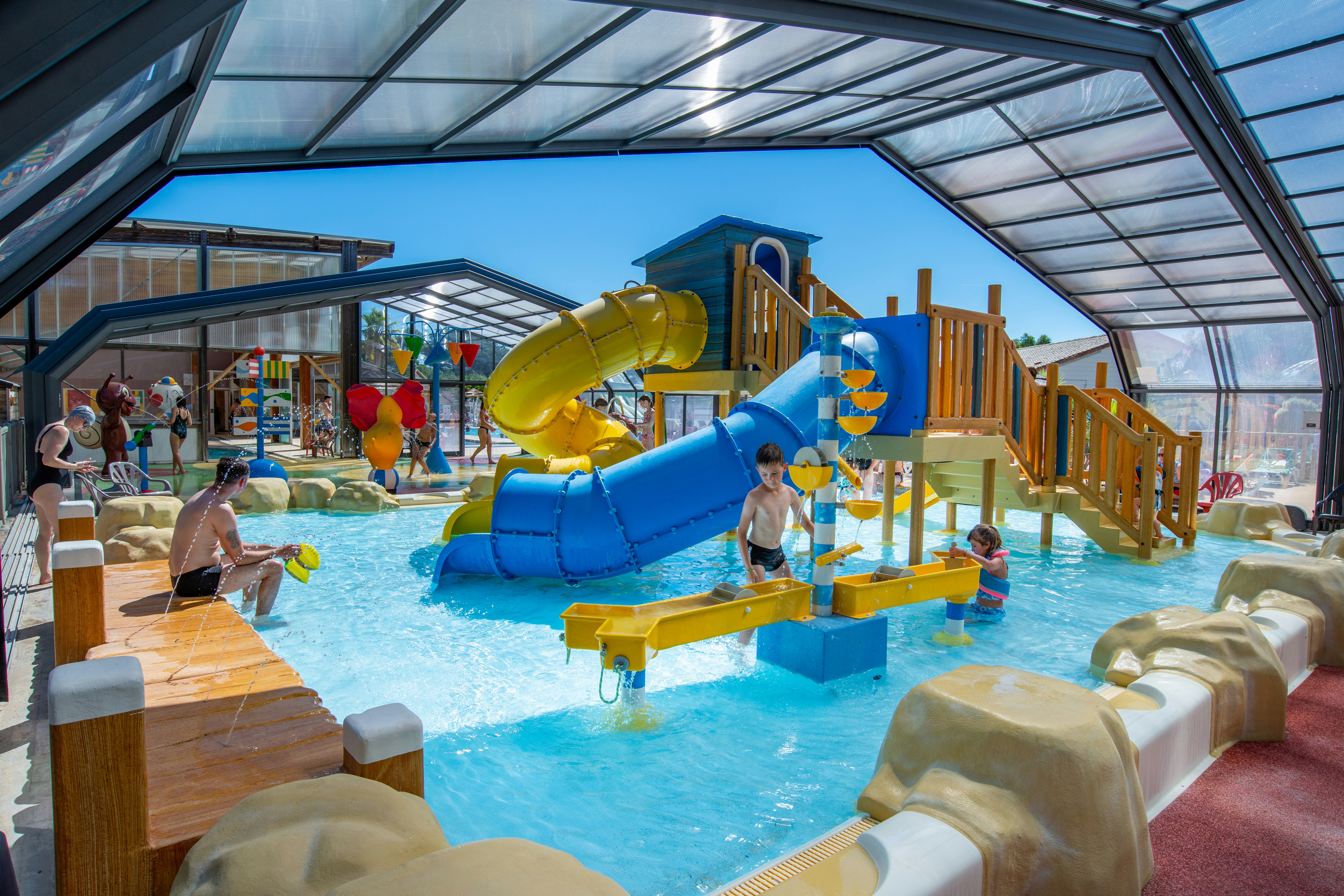 Kinder-Wasserrutsche im überdachten Schwimmbad, mit Holzgeländer, Felsenattrappen und Planschbecken im Hintergrund.