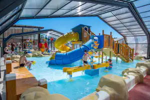 Kinder-Wasserrutsche im überdachten Schwimmbad, mit Holzgeländer, Felsenattrappen und Planschbecken im Hintergrund.