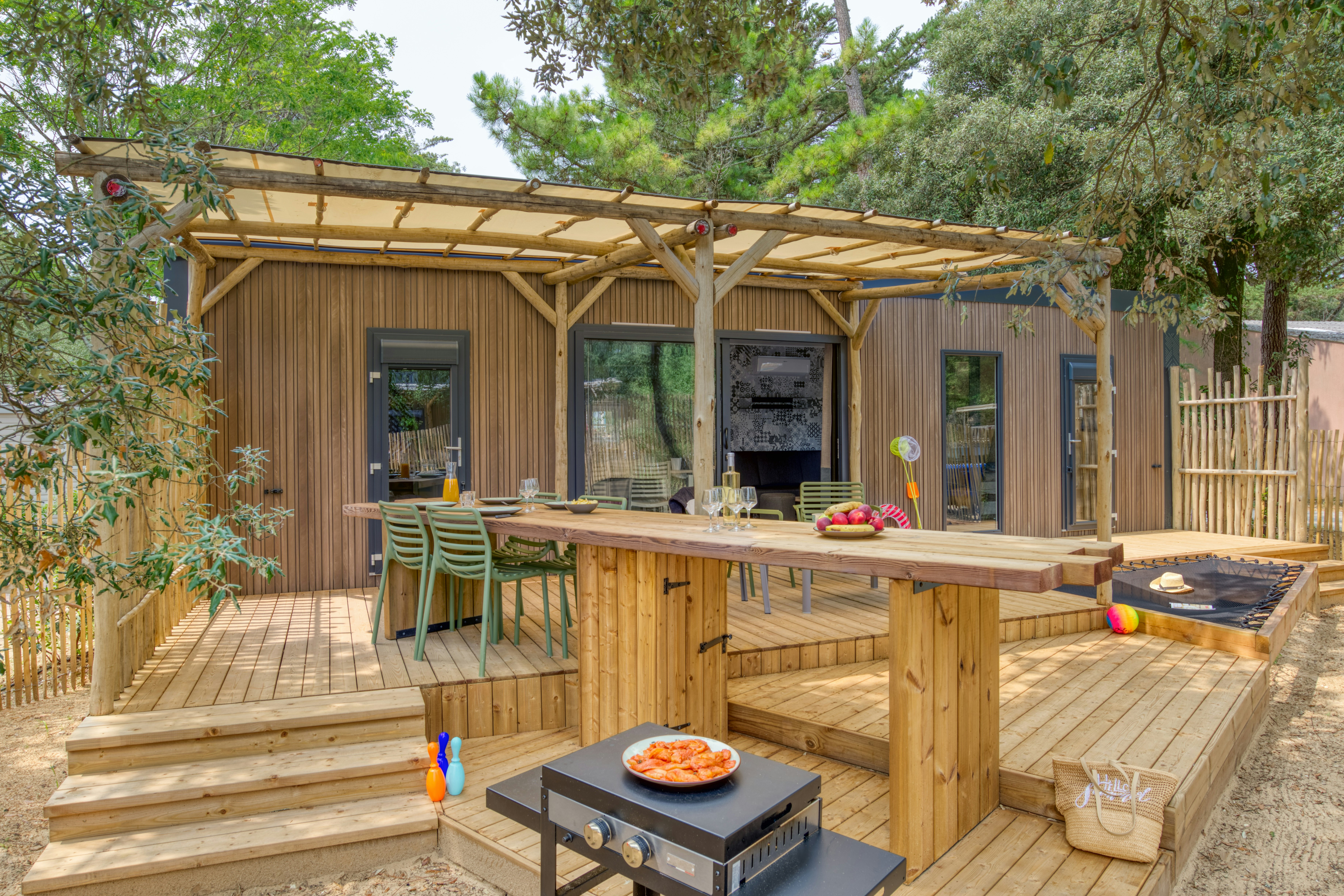 Holzbungalow mit Terrasse, Esstisch und Stühlen, Grill am Boden sowie großen Schiebetüren, umgeben von Bäumen und Holzgeländer.