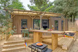 Holzbungalow mit Terrasse, Esstisch und Stühlen, Grill am Boden sowie großen Schiebetüren, umgeben von Bäumen und Holzgeländer.