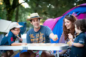 Familie spielt am Camping-Tisch vor bunten Zelten, im Hintergrund Bäume und ein weiteres Zelt.