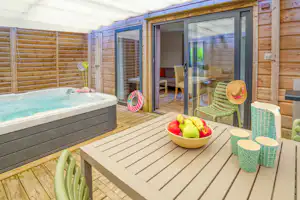 Holzterrasse mit Whirlpool, Glastür zum Wohnbereich und Tisch mit Obstschale, daneben grüne Liegestühle.