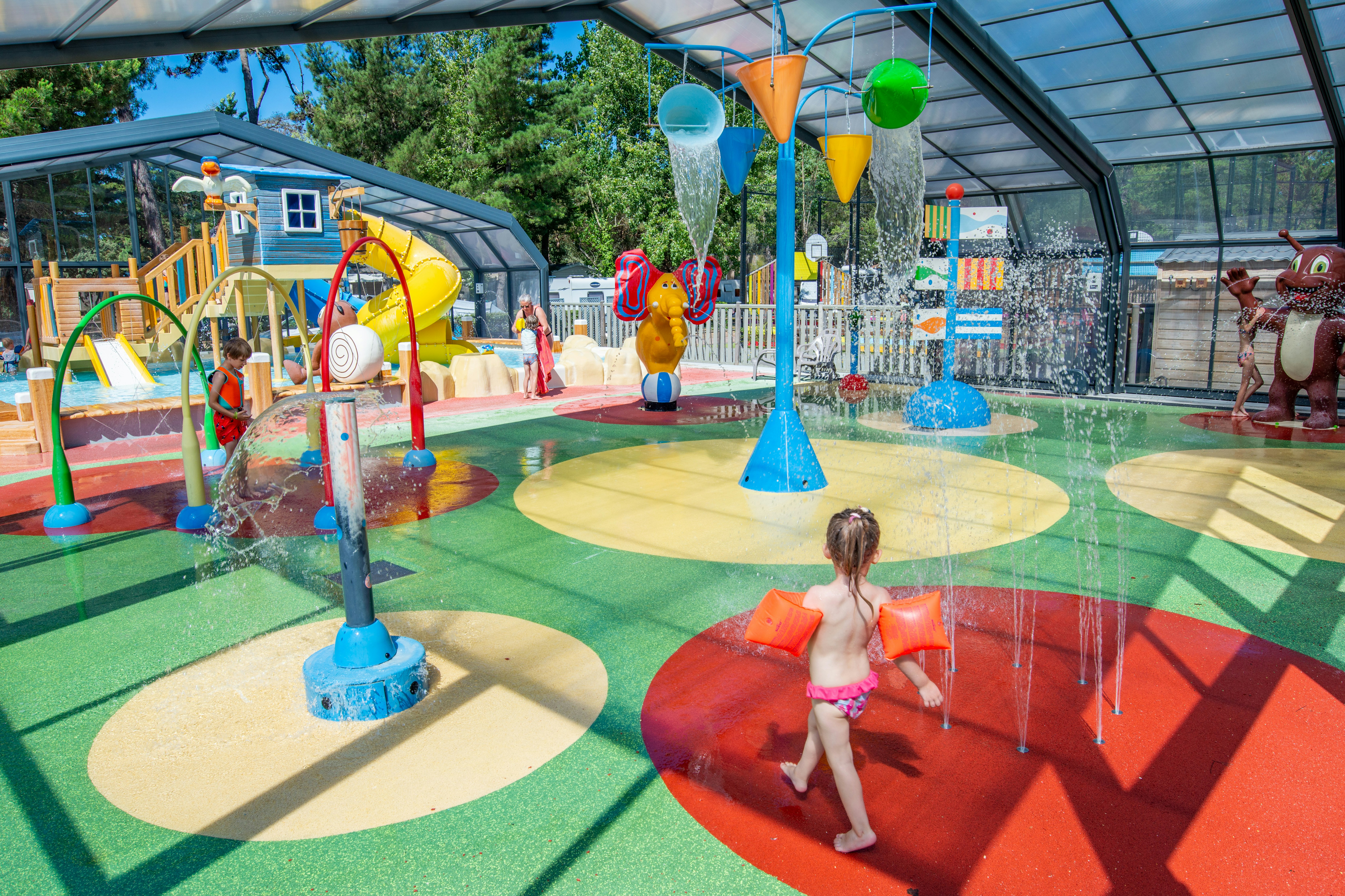 Wasserspielplatz im überdachten Pool mit bunten Spritzfiguren, Wasserfontänen und Kindern auf dem Planschbecken-Boden.