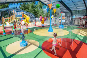 Wasserspielplatz im überdachten Pool mit bunten Spritzfiguren, Wasserfontänen und Kindern auf dem Planschbecken-Boden.