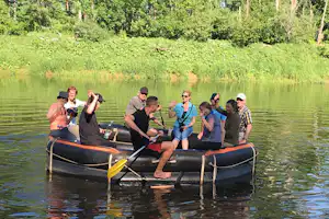 Gruppe im Schlauchboot auf dem See, mit Ruder und Paddel, im Hintergrund dichtes Ufer mit Bäumen.