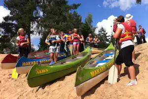 Gruppe mit Schwimmwesten vor mehreren Kanus am sandigen Ufer, im Hintergrund Bäume und blauer Himmel.