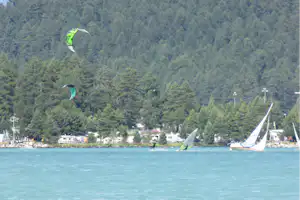 Kites am See vor einem Campingplatz mit Wohnwagen und Bäumen am Ufer, im Hintergrund bewaldete Hügel.