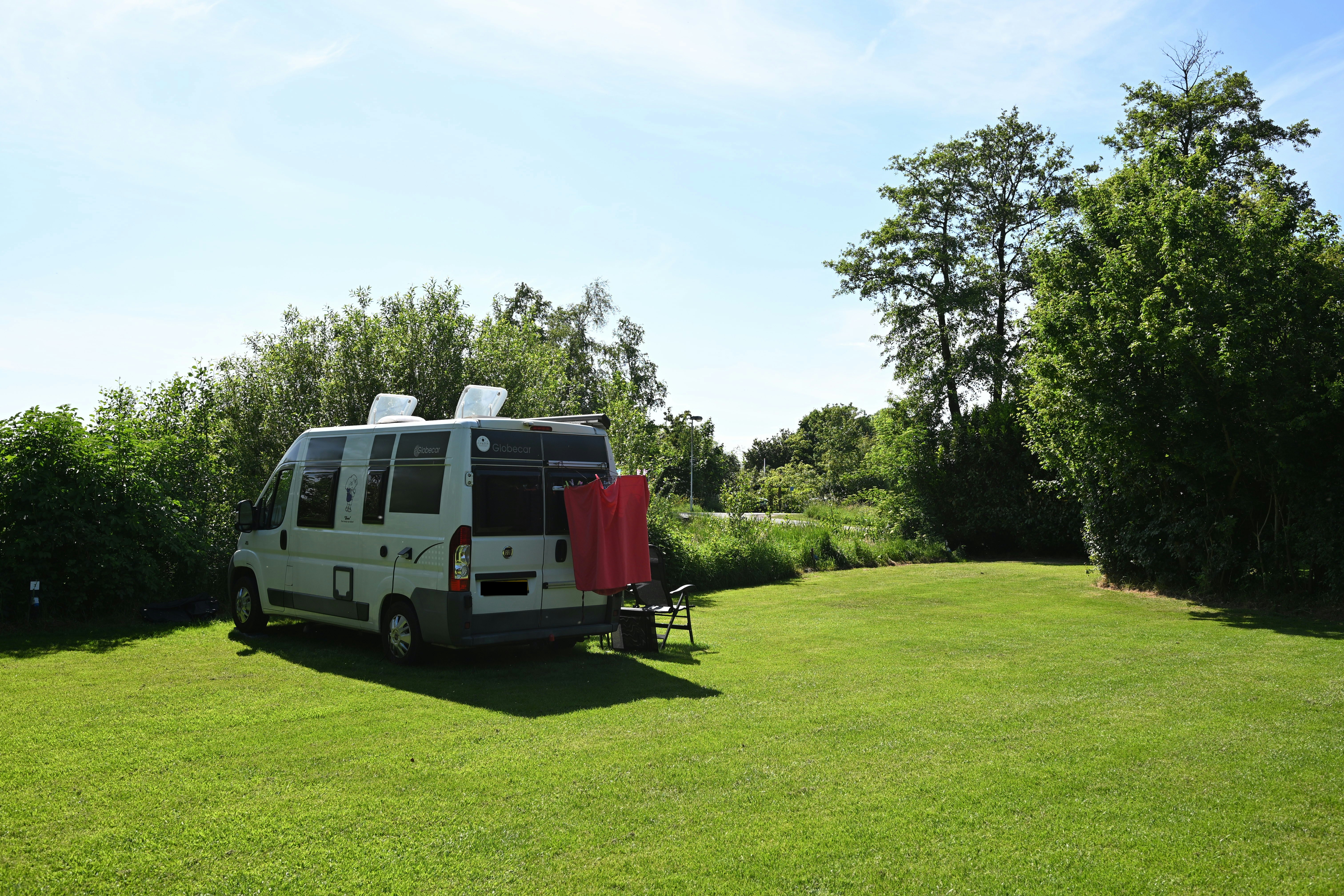 Wohnmobil auf grünem Campingplatz mit geöffnetem Vorzelt, Tisch und Stühlen auf dem Rasen, umgeben von Bäumen.