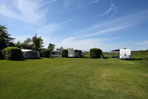 Campingwiese mit mehreren Zelten und Wohnwagen, Hecken und Bäumen am Rand, im Hintergrund ein Wohnmobil.