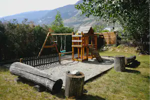 Spielplatz auf dem Campingplatz: Holzschaukel, Rutsche und Sandkasten auf gepflasterter Fläche, umgeben von Bäumen und Bergen.