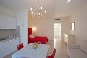 Wohnraum mit rotem Bett, Küchenzeile und Esstisch, Kronleuchter und Flurzugang im hellen Innenbereich.