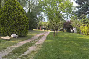 Campingplatz mit Kiesweg, Rasenflächen und Bäumen, im Hintergrund ein Zelt mit rotem Sonnendach.