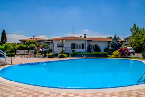 Großer Camping-Pool vor einem weißen Gebäude mit Terrasse, Gartenpflanzen und Sonnenschirm, im Hintergrund weitere Häuser und Autos.