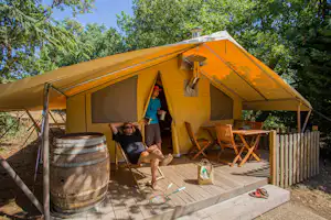 Gelbes Zelt auf einer Holzterrasse mit Sitzgarnitur, Fass und Sonnenschirm im Grünen.
