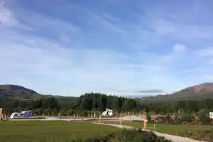Wohnmobile auf einem Campingplatz mit Kieswegen, grünen Wiesen und hohen Bergen im Hintergrund unter blauem Himmel.