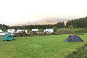 Campingzelt auf grünem Gras, daneben Wohnmobile und weitere Fahrzeuge, im Hintergrund Wald und Berge im Nebel.