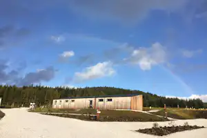 Holzgebäude auf einem Campingplatz, umgeben von Nadelwald, mit Kiesweg und Himmel mit Wolken und Regenbogen.