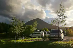 Mehrere Wohnmobile stehen auf einer grünen Campingwiese, im Hintergrund ein Berg und Bäume bei bewölktem Himmel.