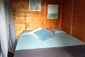 Bett in einer Holzunterkunft mit gestreifter Bettwäsche und Kissen, Vorhang am Fenster und zwei Wandbildern.