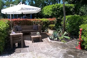 Camping Smeraldo - Sitzmöglichkeit unter Sonnenschirm umgeben von Sträuchern und Bäumen