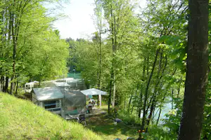 Wohnwagen und Zelte auf einem Campingplatz im Grünen, mit Bäumen, Rasenfläche und Blick auf den See.