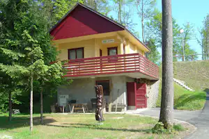 Holzhütte mit roter Dachfläche und Balkon, Sitzbank und Tisch vor dem Eingang, umgeben von Bäumen und Rasen.