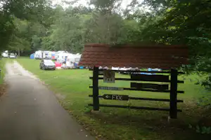 Wegweiser mit FKK-Schildern am Campingplatz, daneben Wohnwagen und Autos auf dem Grasweg unter Bäumen.