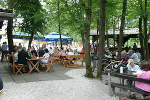 Biergarten am Campingplatz mit langen Holztischen, vielen Gästen und Fahrrädern zwischen Bäumen im Schatten.