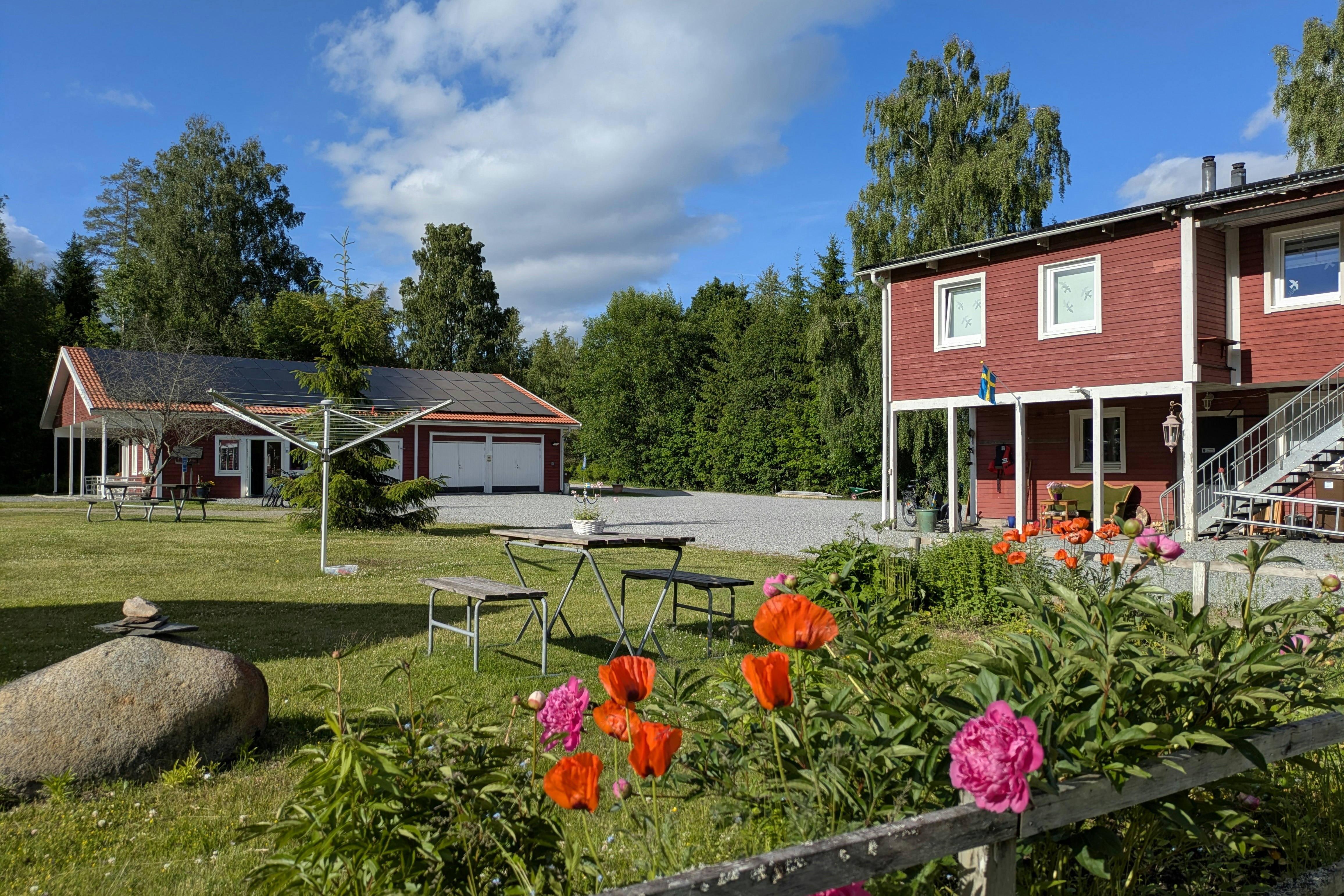 Blumenbeet mit roten und pinken Blüten vor einem Campingplatz, daneben Rasen und Gartenmöbel vor roten Ferienhäusern.