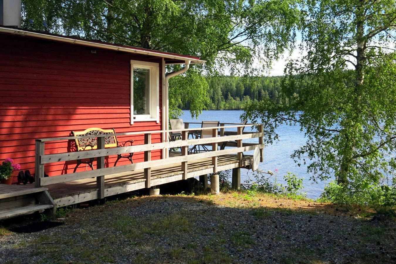 Rote Holzunterkunft mit Veranda und Geländer am Campingplatz, Blick auf den See und Bäume im Hintergrund.