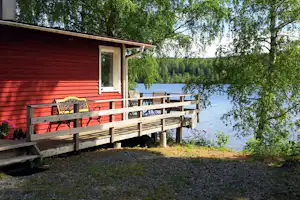 Rote Holzunterkunft mit Veranda und Geländer am Campingplatz, Blick auf den See und Bäume im Hintergrund.