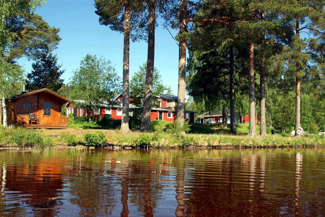 Holzhäuser und Wohnwagen am Ufer eines Sees, mit Bäumen im Hintergrund und ruhigem Wasser vor dem Campingplatz.