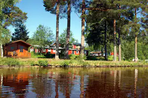 Holzhäuser und Wohnwagen am Ufer eines Sees, mit Bäumen im Hintergrund und ruhigem Wasser vor dem Campingplatz.